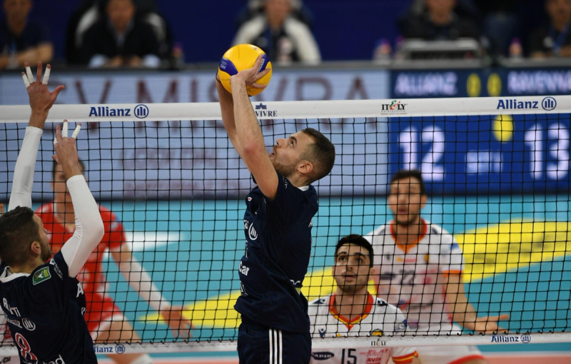 A volleyball player in a dark blue uniform jumps to set the ball near the net, while opposing players in white and orange uniforms prepare to respond. The game takes place in an indoor arena with spectators in the background.