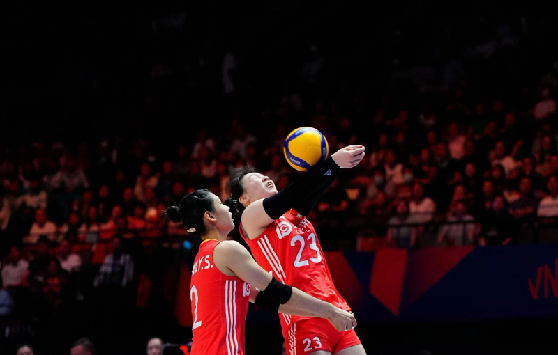 Two volleyball players in red uniforms are attempting to hit a ball mid-air during a match, with a blurred audience in the background.