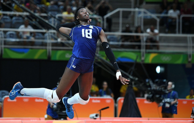 A volleyball player in a blue uniform, wearing the number 18, is airborne during a jump serve in an indoor arena. The player looks focused and athletic, with spectators visible in the background.