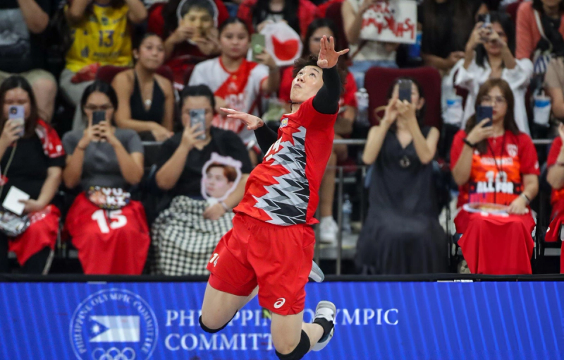 A volleyball player in a red uniform jumps to hit the ball during a game. Spectators in the background capture the moment on their phones, some holding posters and wearing team colors.