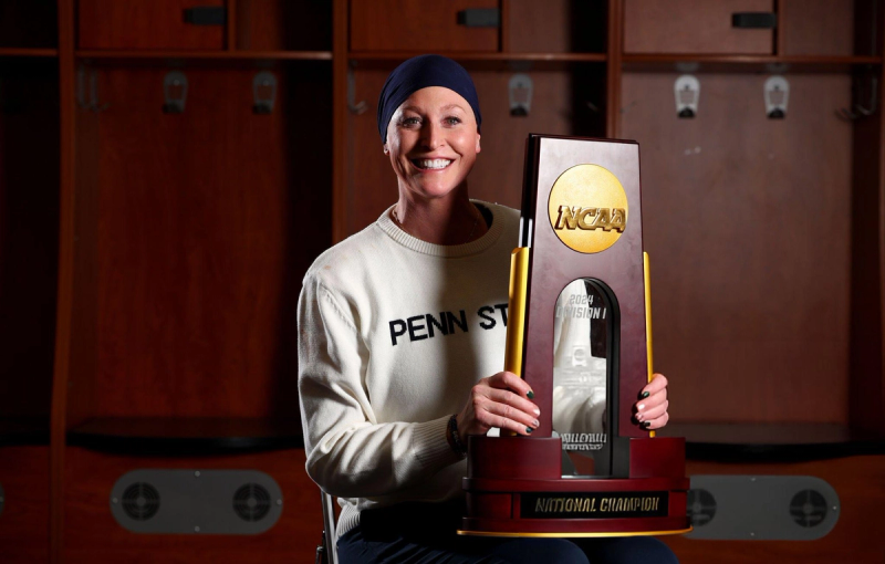 A person wearing a Penn State sweater and headband smiles while holding an NCAA 2024 Division I National Champion trophy in a locker room.