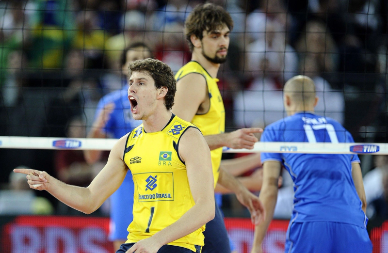 A volleyball player in a yellow jersey energetically points and reacts during a match. Another teammate in yellow and an opponent in blue are visible in the background near the net.