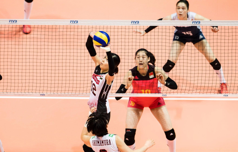 Two volleyball players are at the net during a match. One player is jumping to set the ball while the other is preparing to block. The action takes place on a court with a neutral-colored floor.