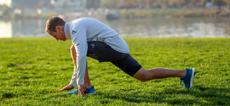 A person in sports attire is stretching in a park, lunging forward with one knee bent and the other leg extended. There is a river and blurred trees in the background.