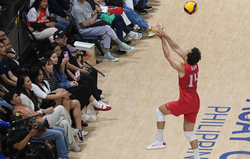 Volleyball player in red gear is jumping to reach a ball near the court's edge, with an audience closely watching.