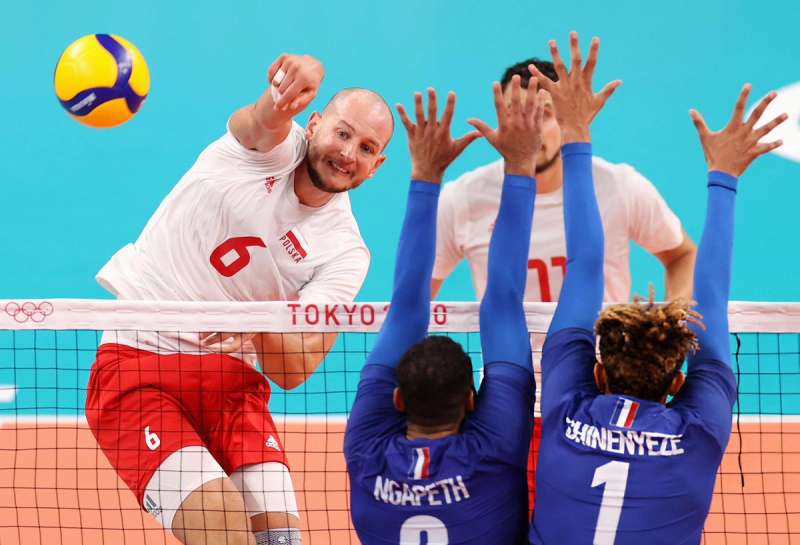 A volleyball player in a white and red uniform spikes the ball over the net while two opposing players in blue jerseys attempt to block. The net displays "Tokyo 2020."