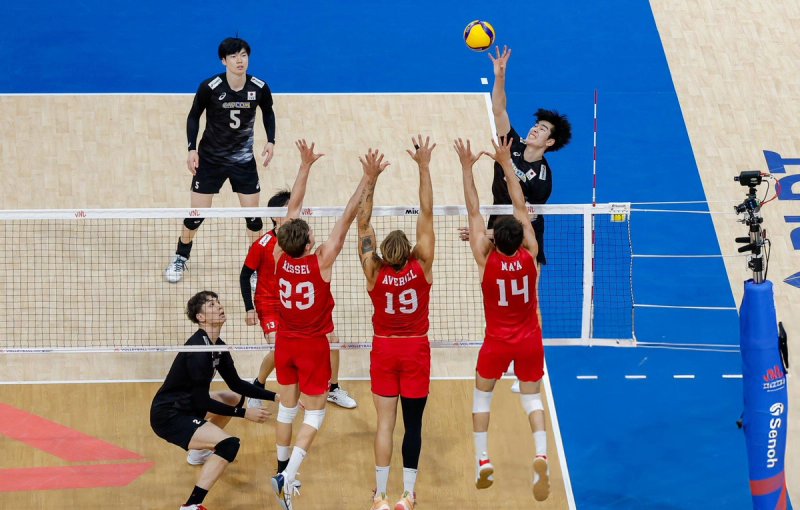 A volleyball player in a black uniform is spiking the ball over the net as three opposing players in red uniforms jump to block, with another player in black crouched nearby on a wooden court.