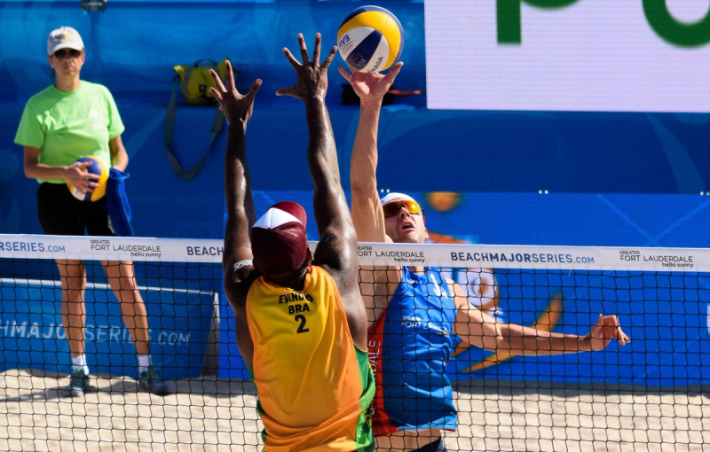 Two beach volleyball players compete at the net, with one player in a yellow jersey attempting to block a ball hit by a player in a blue jersey. An official stands in the background holding another ball.