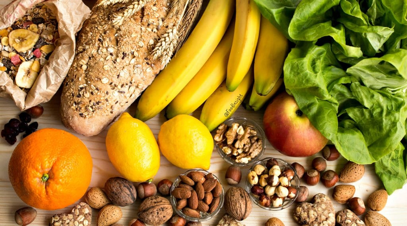 A selection of fresh produce and nuts: bananas, a loaf of multigrain bread, leafy greens, a red apple, lemons, an orange, mixed nuts, and a bag of muesli.