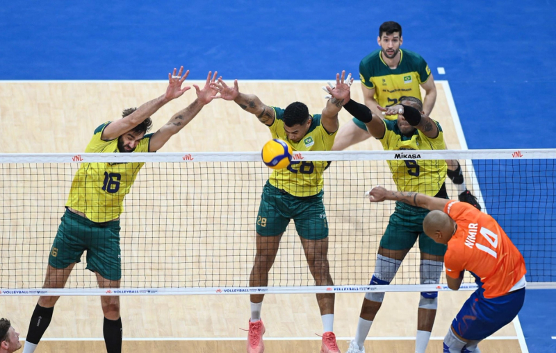 Men's volleyball game in action, with three players in yellow jerseys attempting to block a player in an orange jersey at the net.
