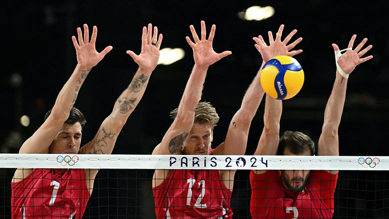 Three volleyball players in red jerseys attempt to block a ball over the net, with "Paris 2024" and Olympic rings visible.