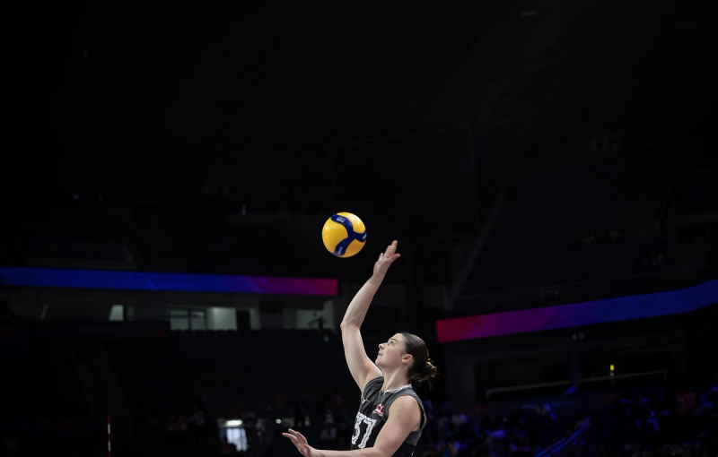 A volleyball player in a dark uniform jumps to hit a volleyball in mid-air in a dimly lit arena.