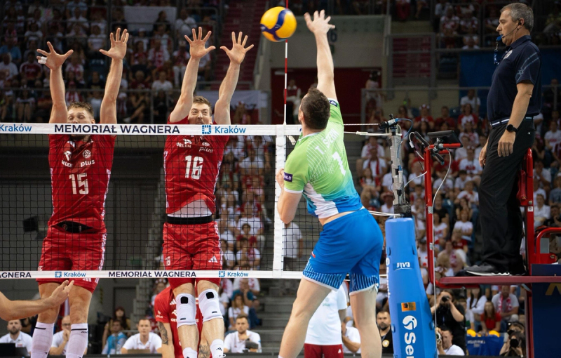 Volleyball match action with a player in a green and blue uniform spiking the ball over the net, while two opposing players in red uniforms attempt to block him. An official stands to the side on a raised platform, overseeing the game. The arena is filled with spectators.