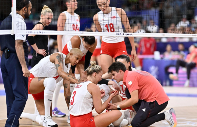 A group of volleyball players and a coach attend to a teammate sitting on the floor, comforting her during a break in the match.