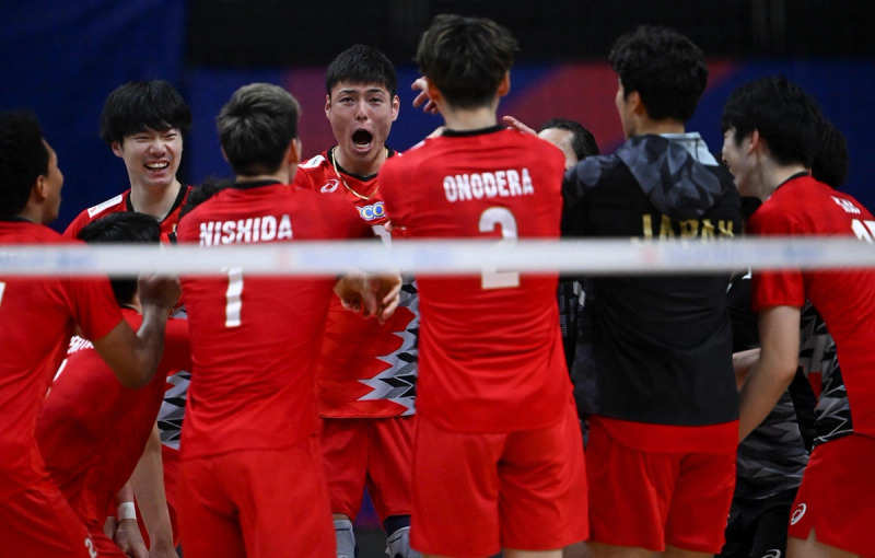 A group of male volleyball players in red uniforms celebrate energetically on the court. They are smiling, laughing, and appear joyful, with visible team camaraderie.