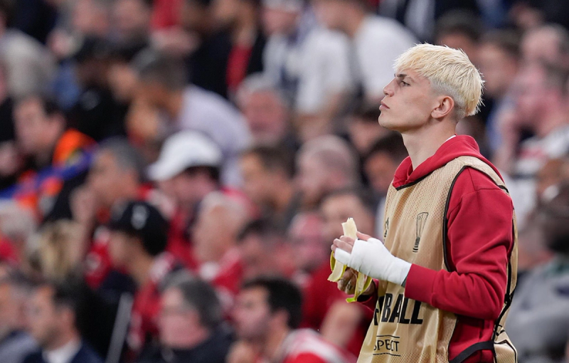 A person with short blond hair, wearing a red hoodie and a tan bib, stands in front of a blurred crowd, eating a banana. Their left hand is bandaged.