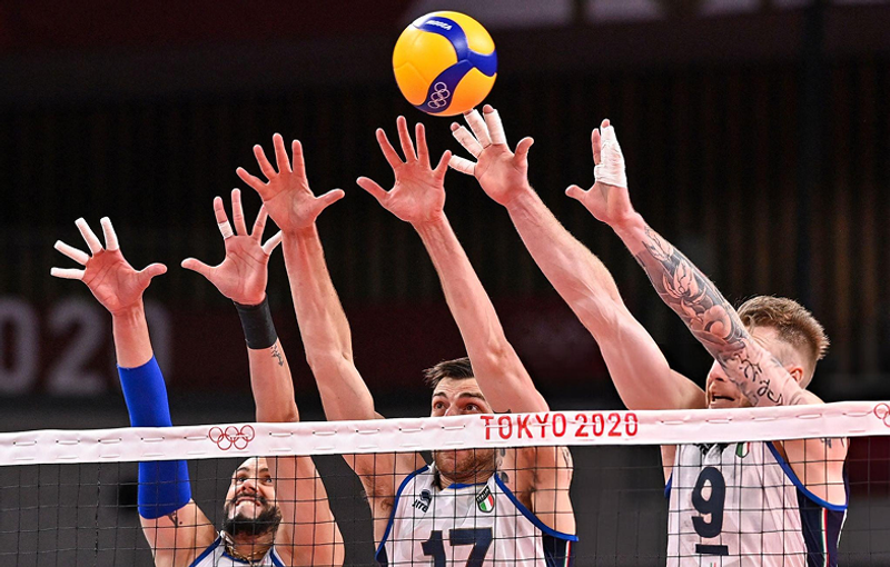 Volleyball players jumping to block a ball over the net during the Tokyo 2020 Olympics match.