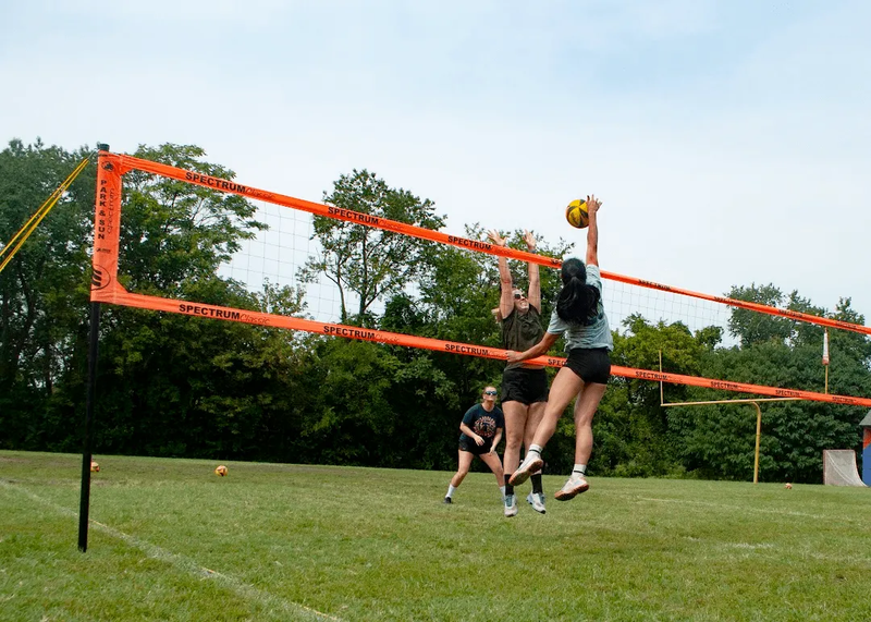 Three people playing volleyball on a grassy field. One player is jumping to hit the ball, with the net in the foreground and trees in the background.