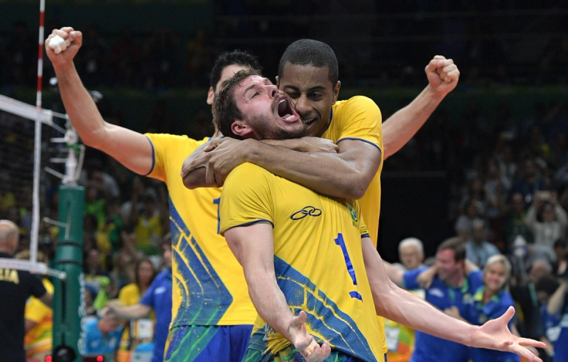Volleyball players in yellow jerseys celebrating joyfully on the court. One player is embracing another with visible excitement, while another player raises a fist in triumph.