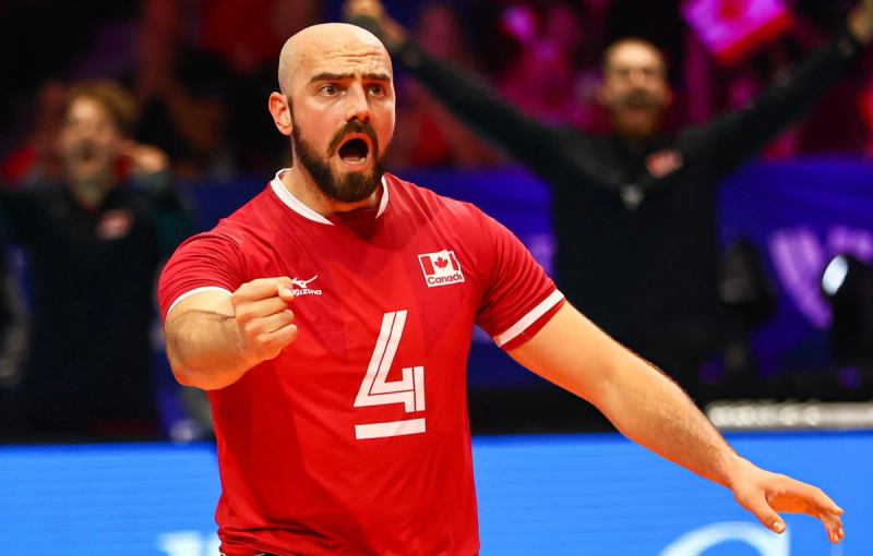 A volleyball player wearing a red jersey with the number 4 and a Canadian flag, looks energized and determined during a match, with a blurred crowd in the background.