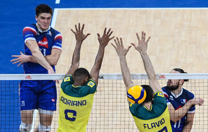 A volleyball player in a blue jersey is attacking at the net, while two players in yellow jerseys attempt to block the ball.