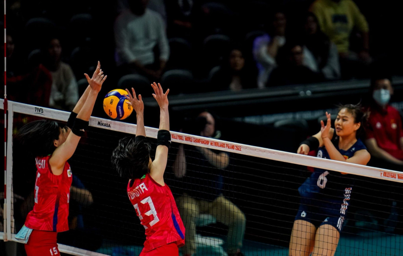 A volleyball player in a blue jersey spikes the ball towards two players in red jerseys attempting to block it at the net during a match.