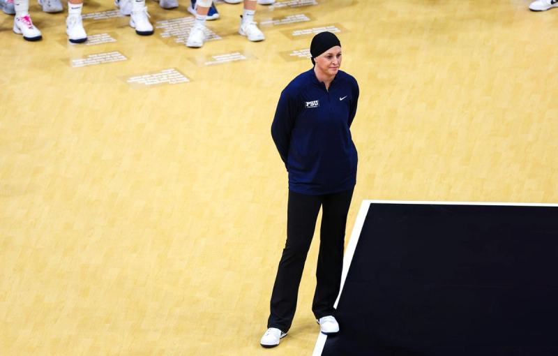 A person in a navy sports jacket and black pants stands on a gym court with hands behind their back. White text is visible on the floor, with people in athletic shoes standing in the background.