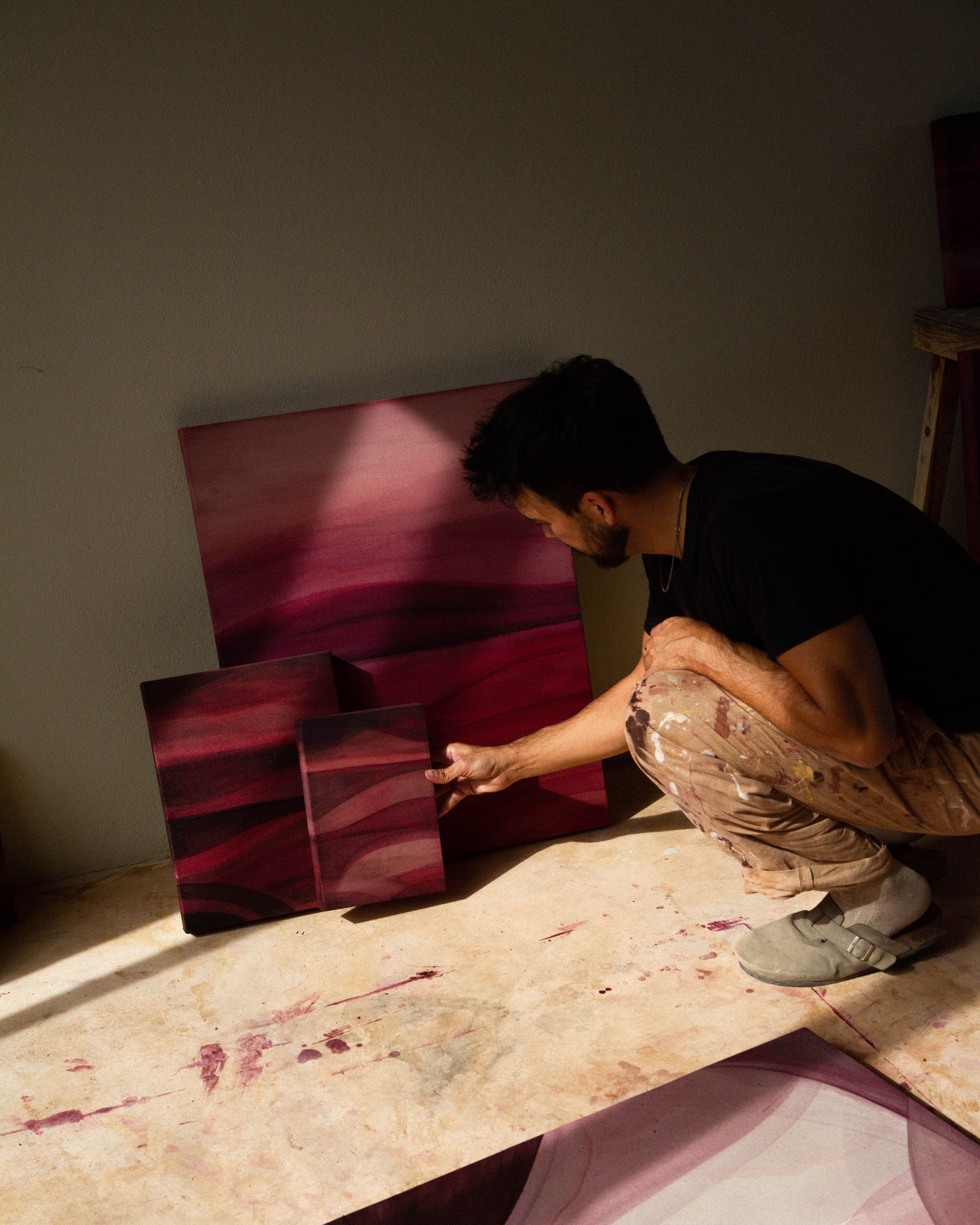 An artist crouching in a studio examining an abstract painting beside other works on the floor.