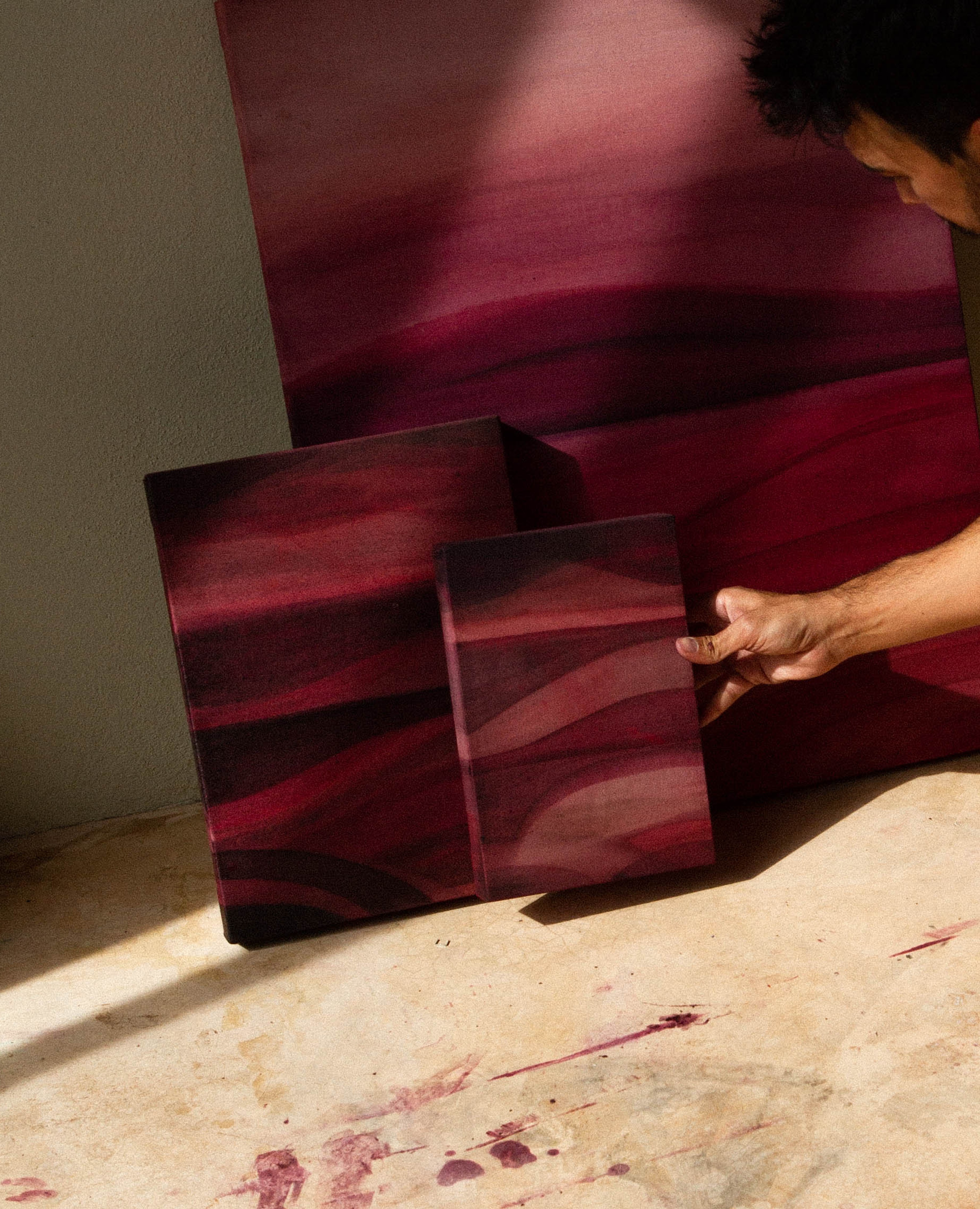 An artist crouching in a studio examining an abstract painting beside other works on the floor.