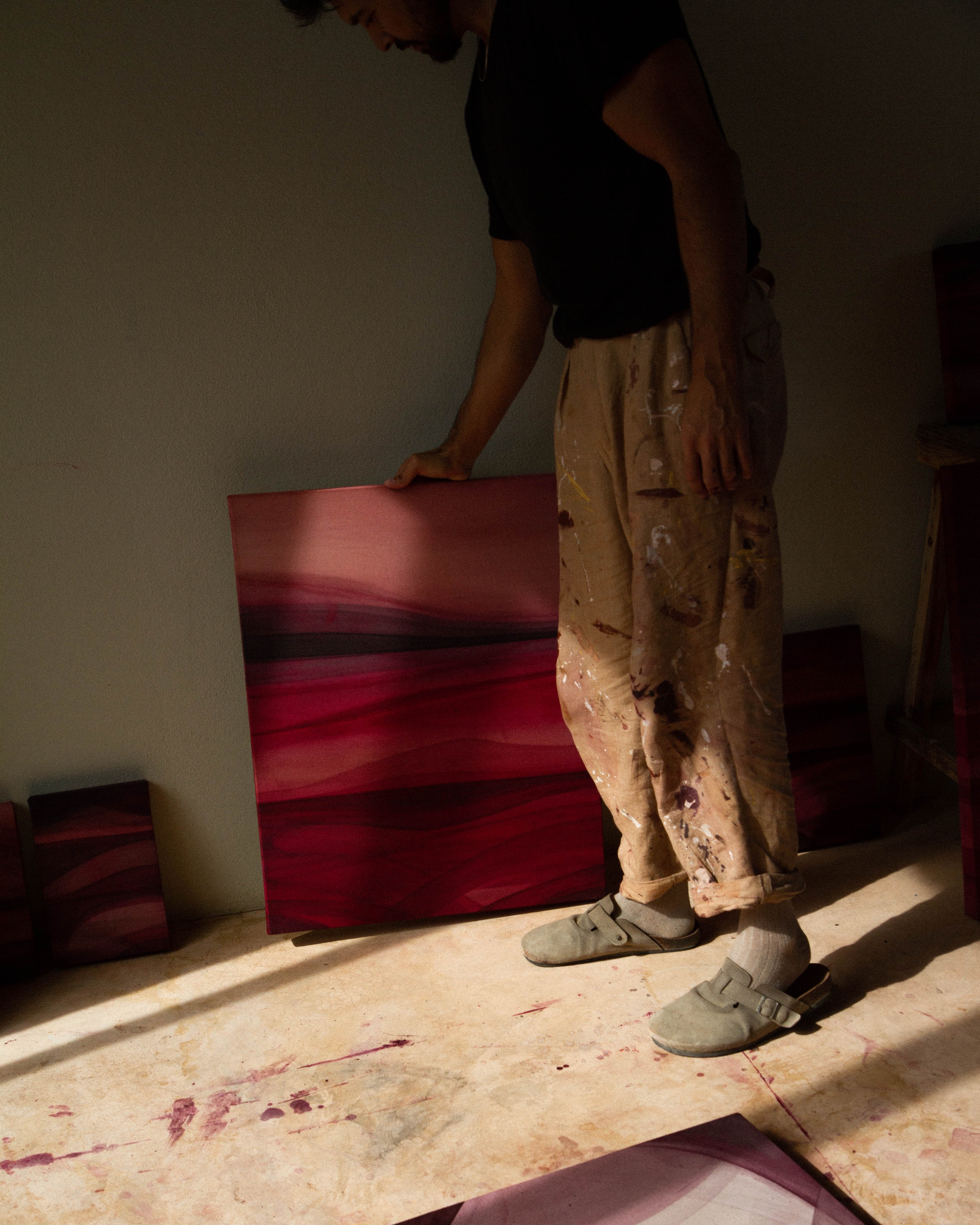 An artist standing in a studio holding an abstract painting beside other works in natural light.