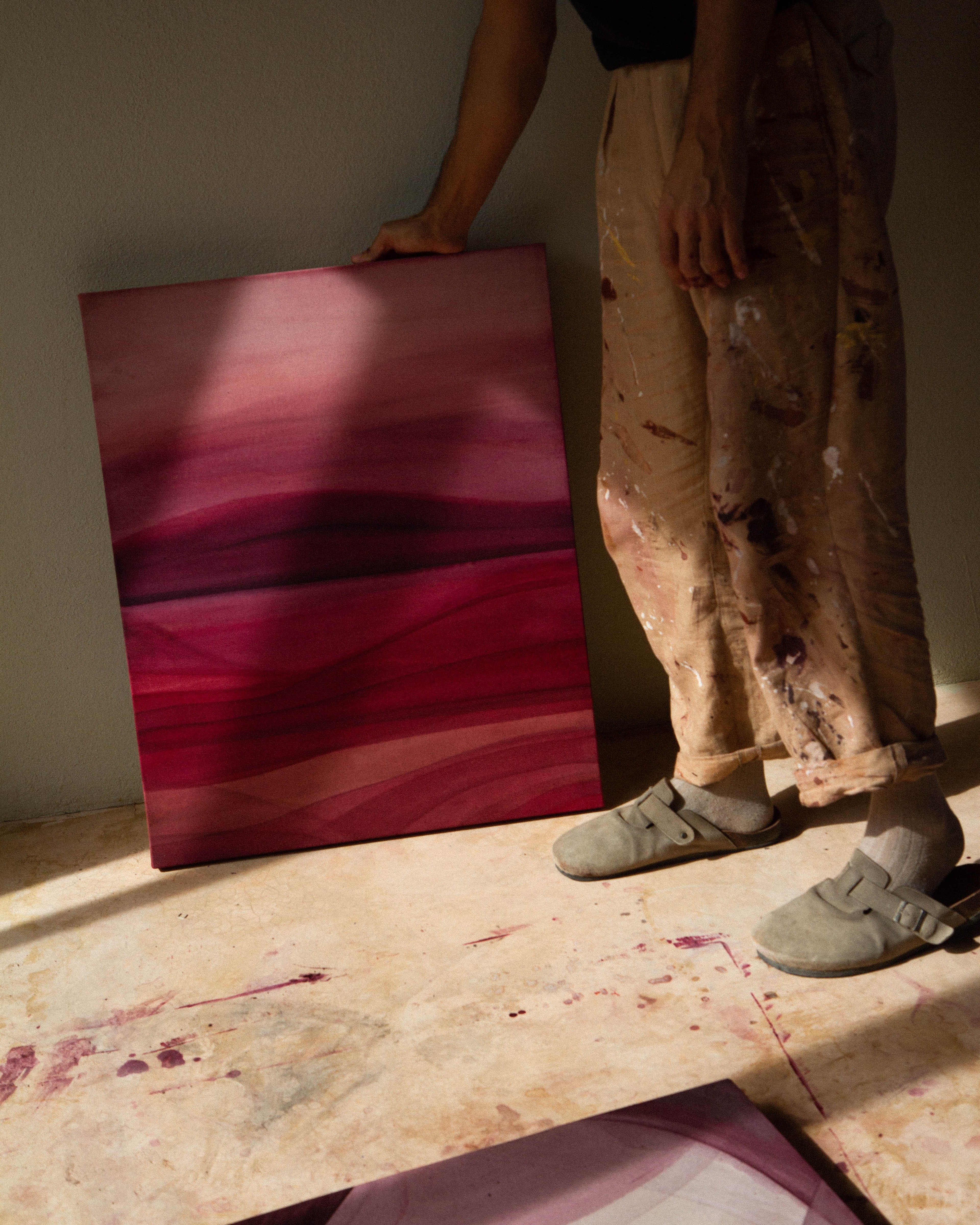An artist standing in a studio holding an abstract painting beside other works in natural light.