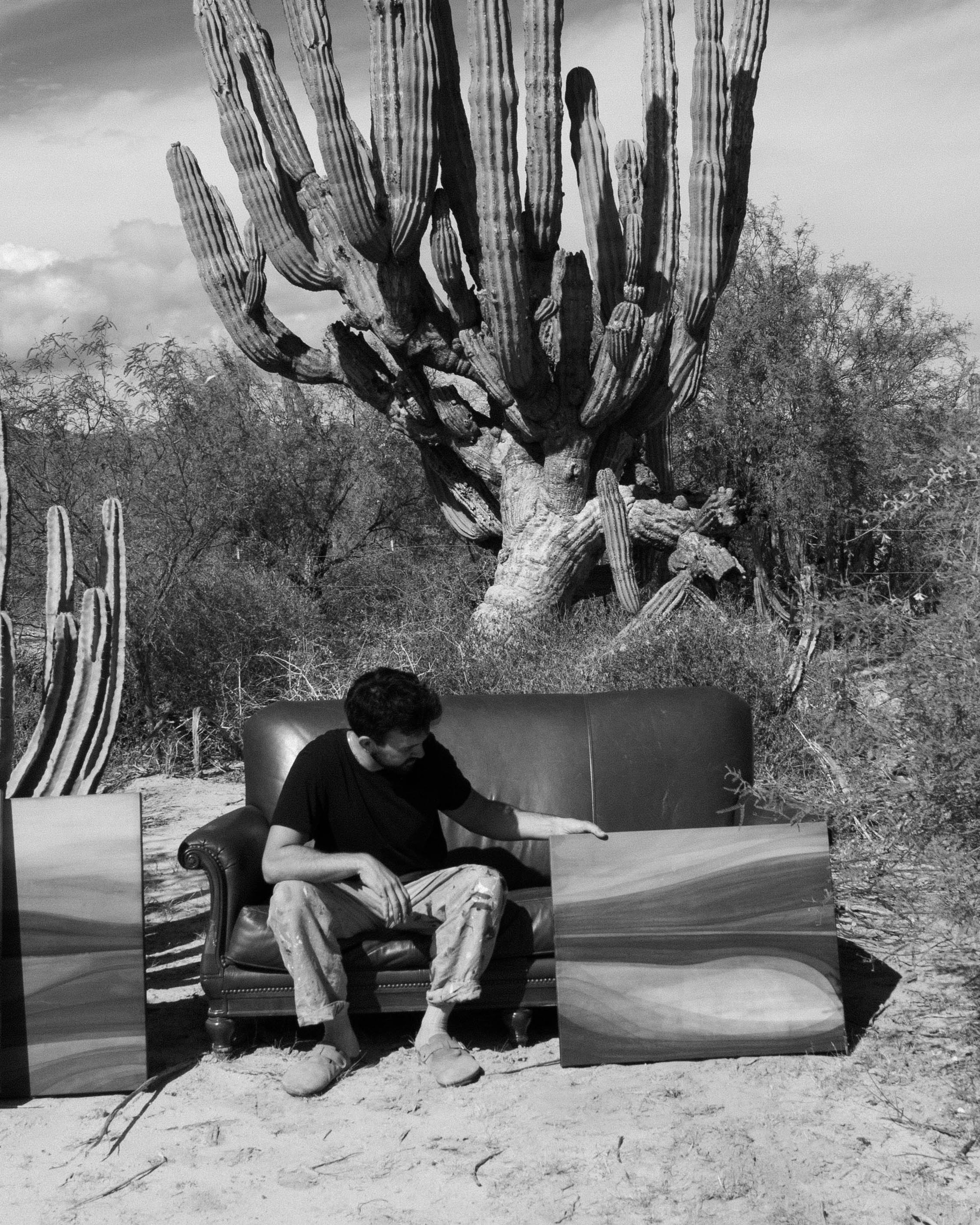 An artist seated outdoors in a desert landscape holding an abstract painting with cactus plants in the background.