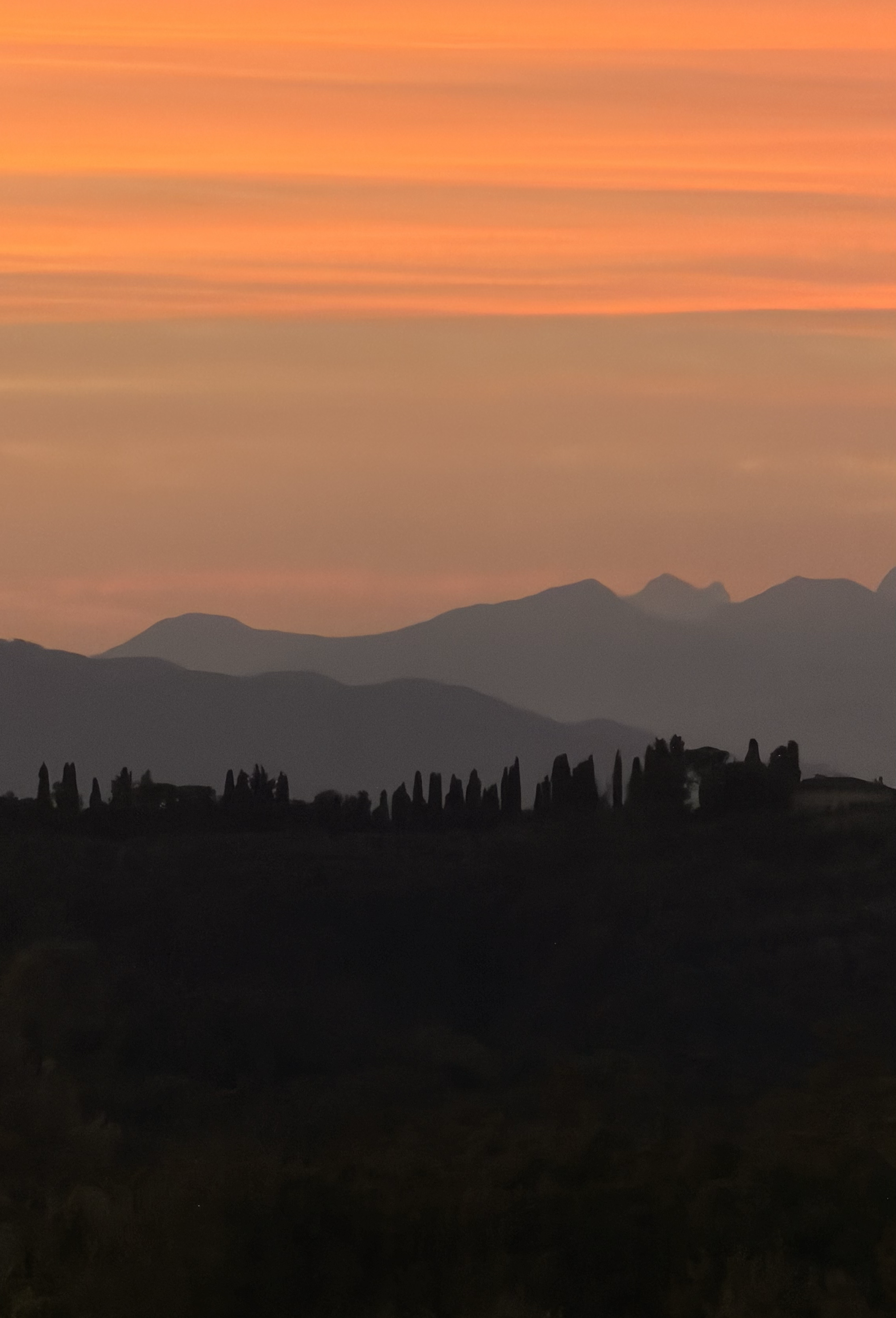 Orange sunset over the Tuscan hills, with cypress trees silhouetted on the horizon, seen from Villa Lena.