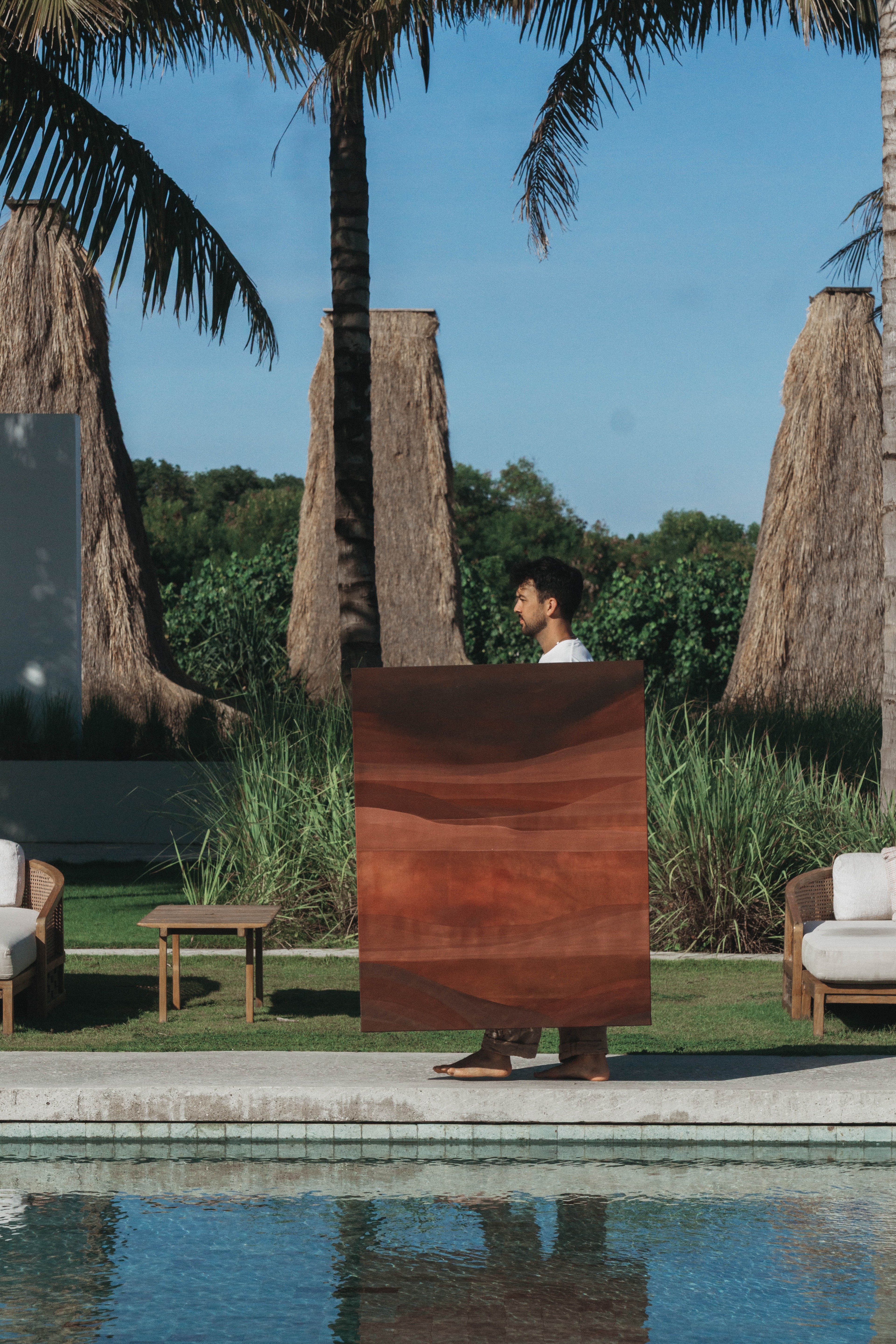 An artist holding a large abstract painting outdoors beside a pool with trees in the background.