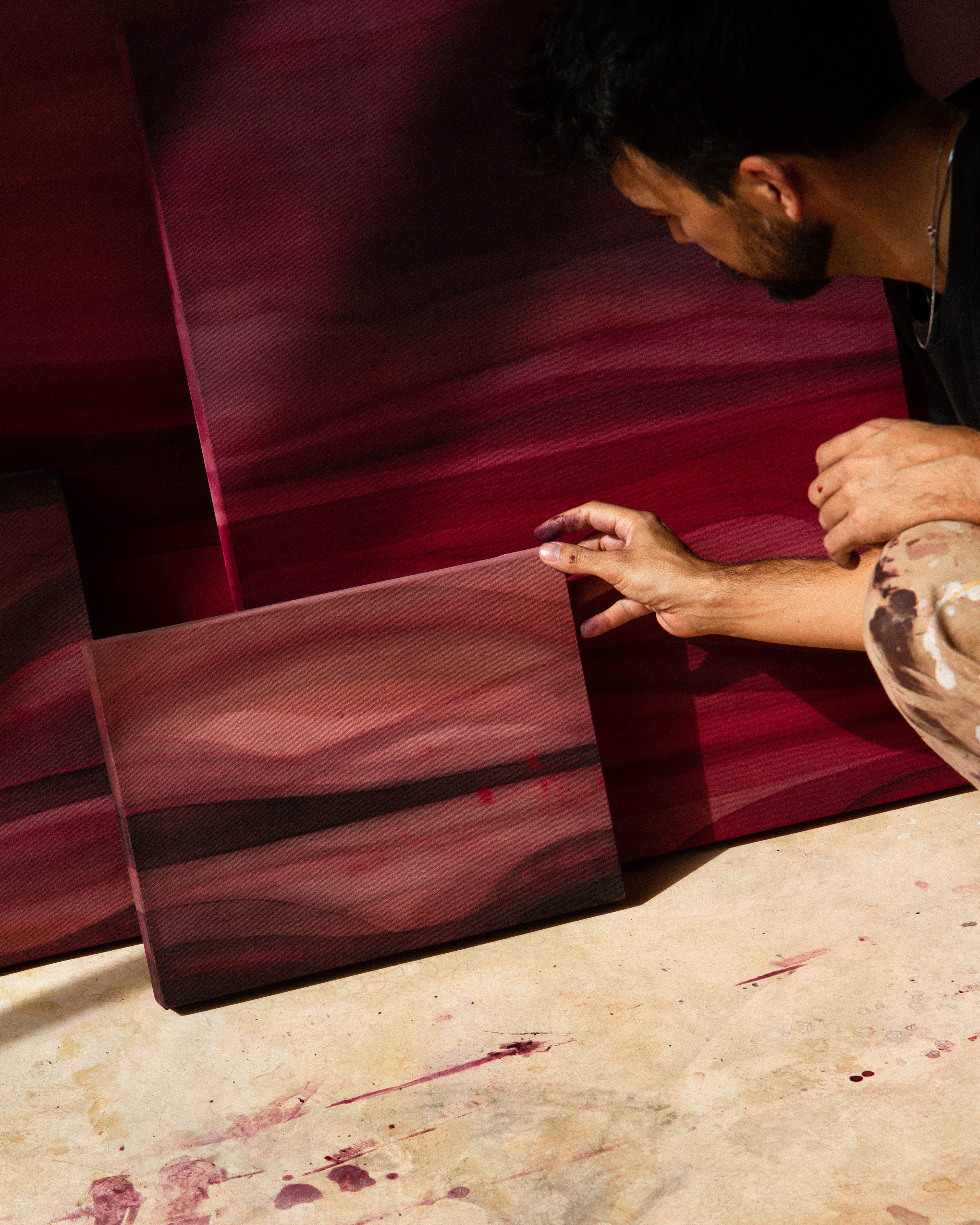An artist crouching in a studio examining an abstract painting beside other works on the floor.
