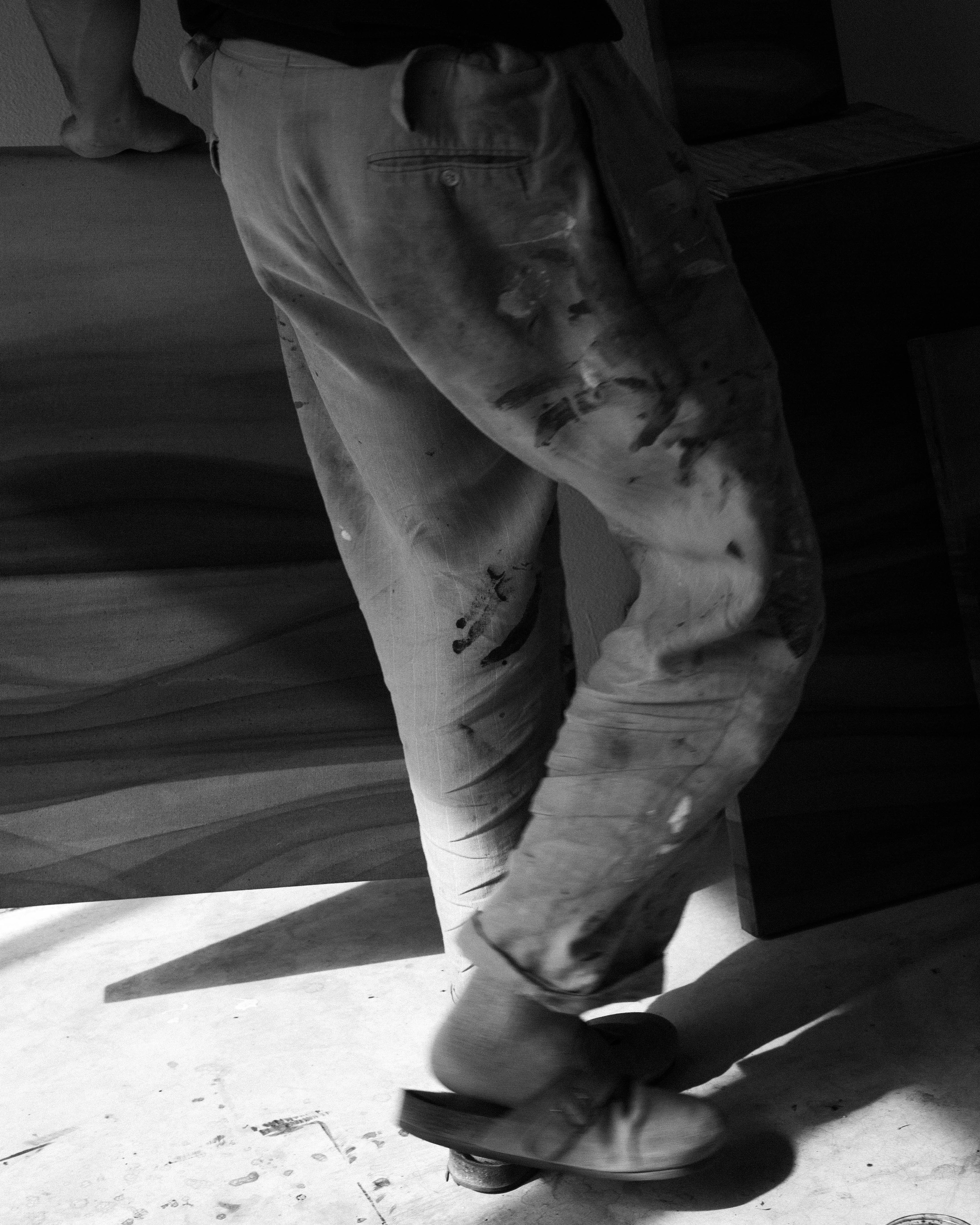 An artist standing in a studio holding an abstract painting beside other works in natural light.