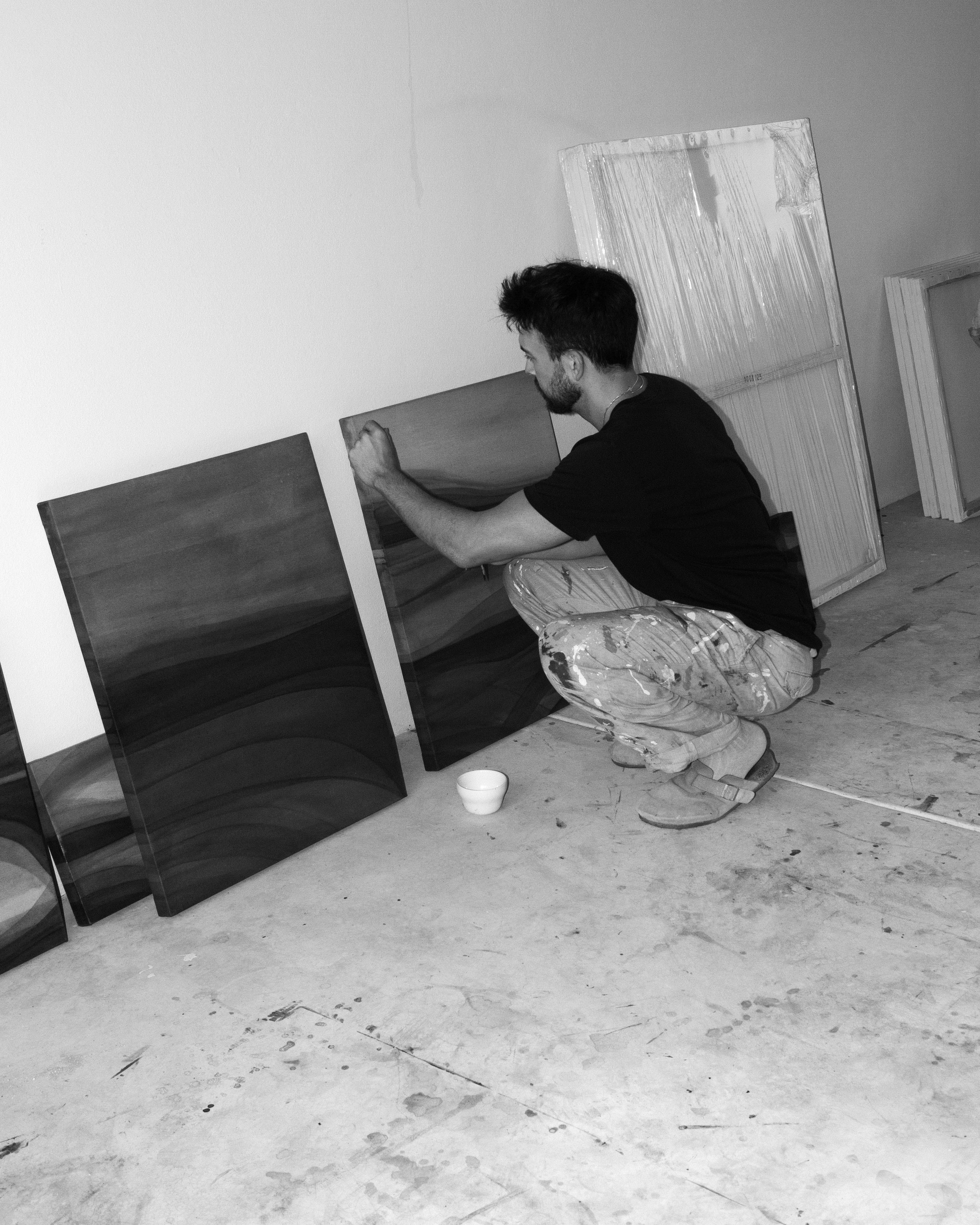 An artist crouching in a studio while working on abstract paintings placed on the floor in natural light.