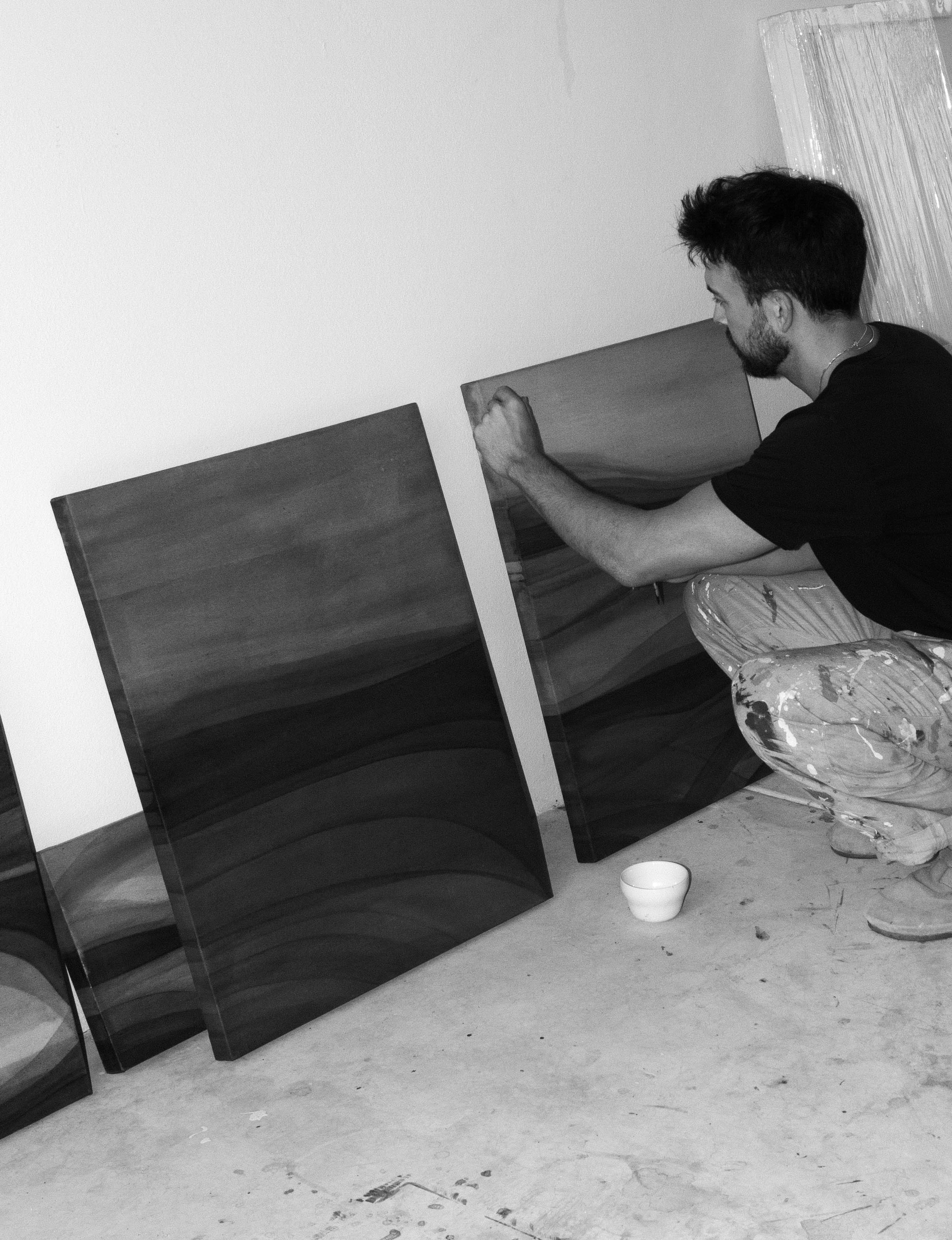 An artist crouching in a studio while working on abstract paintings placed on the floor in natural light.