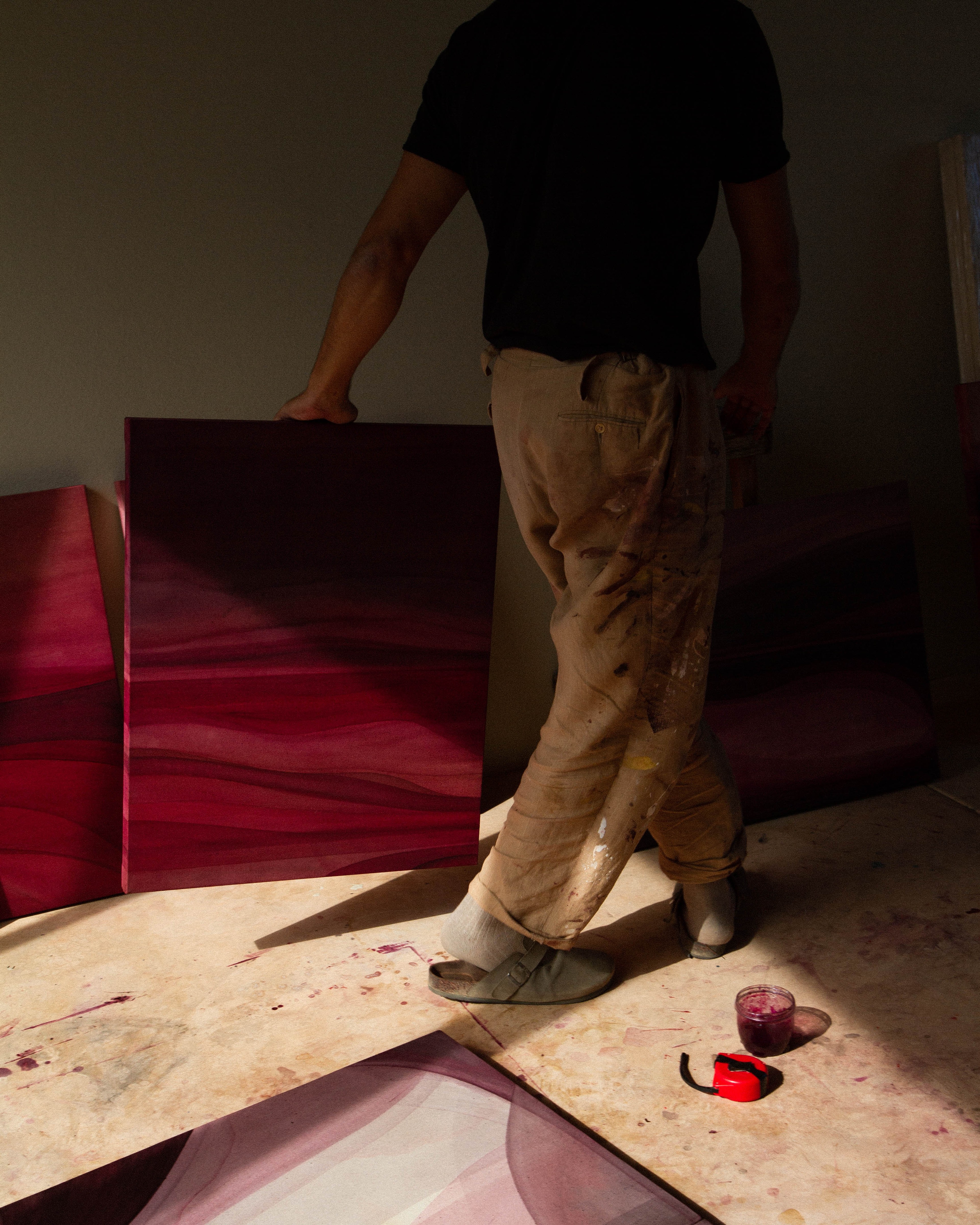 An artist standing in a studio holding an abstract painting beside other works in natural light.