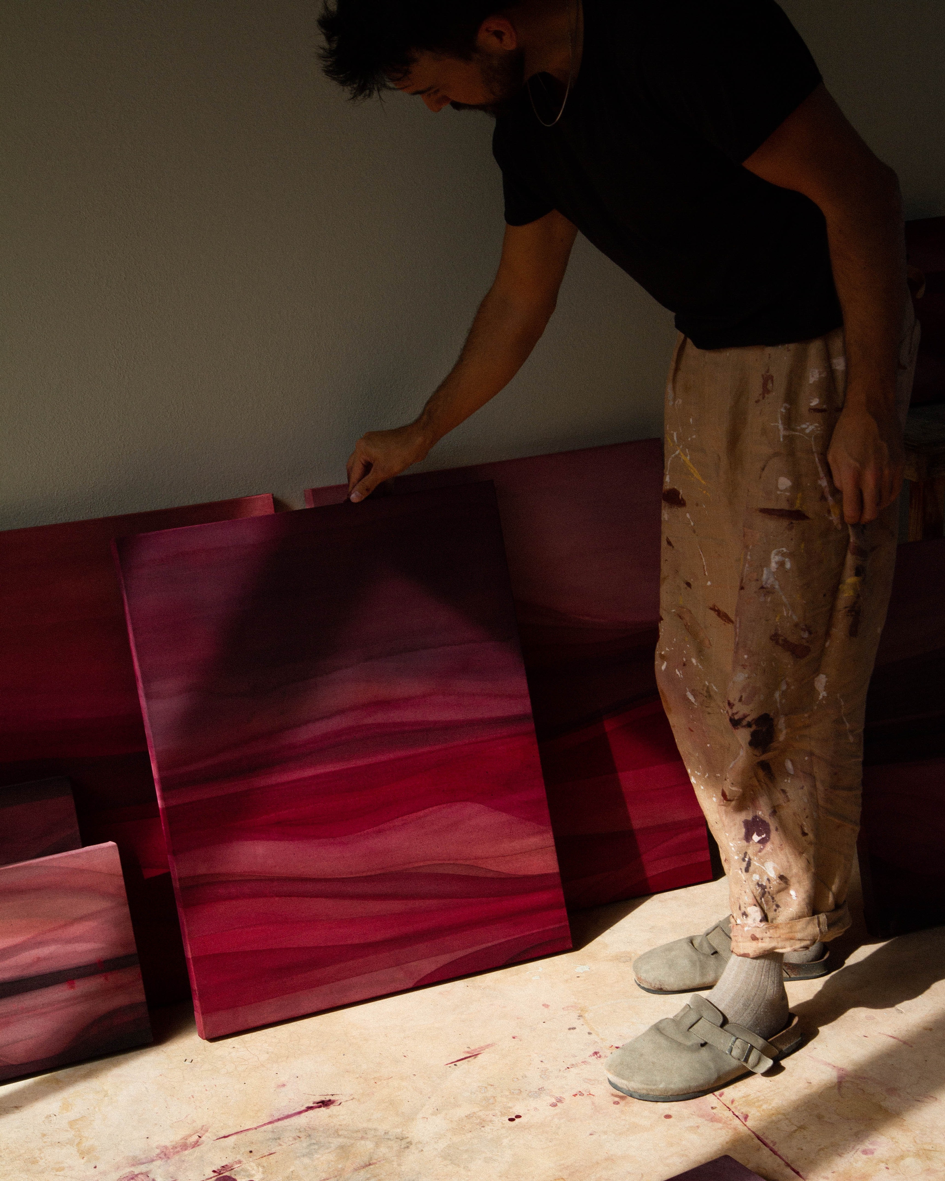 An artist standing in a studio holding an abstract painting beside other works in natural light.