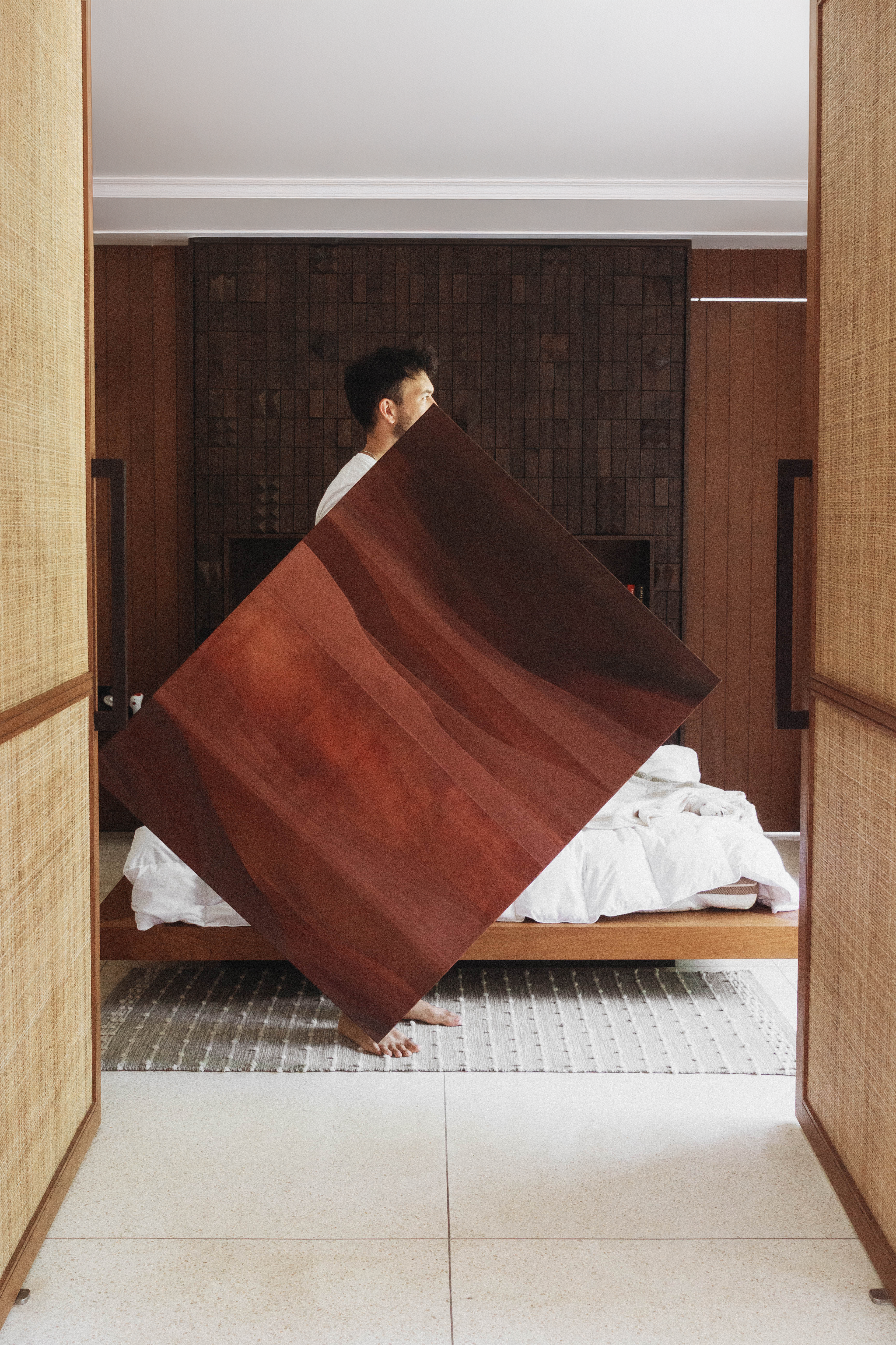 An artist holding a large abstract painting inside an interior space with natural light.