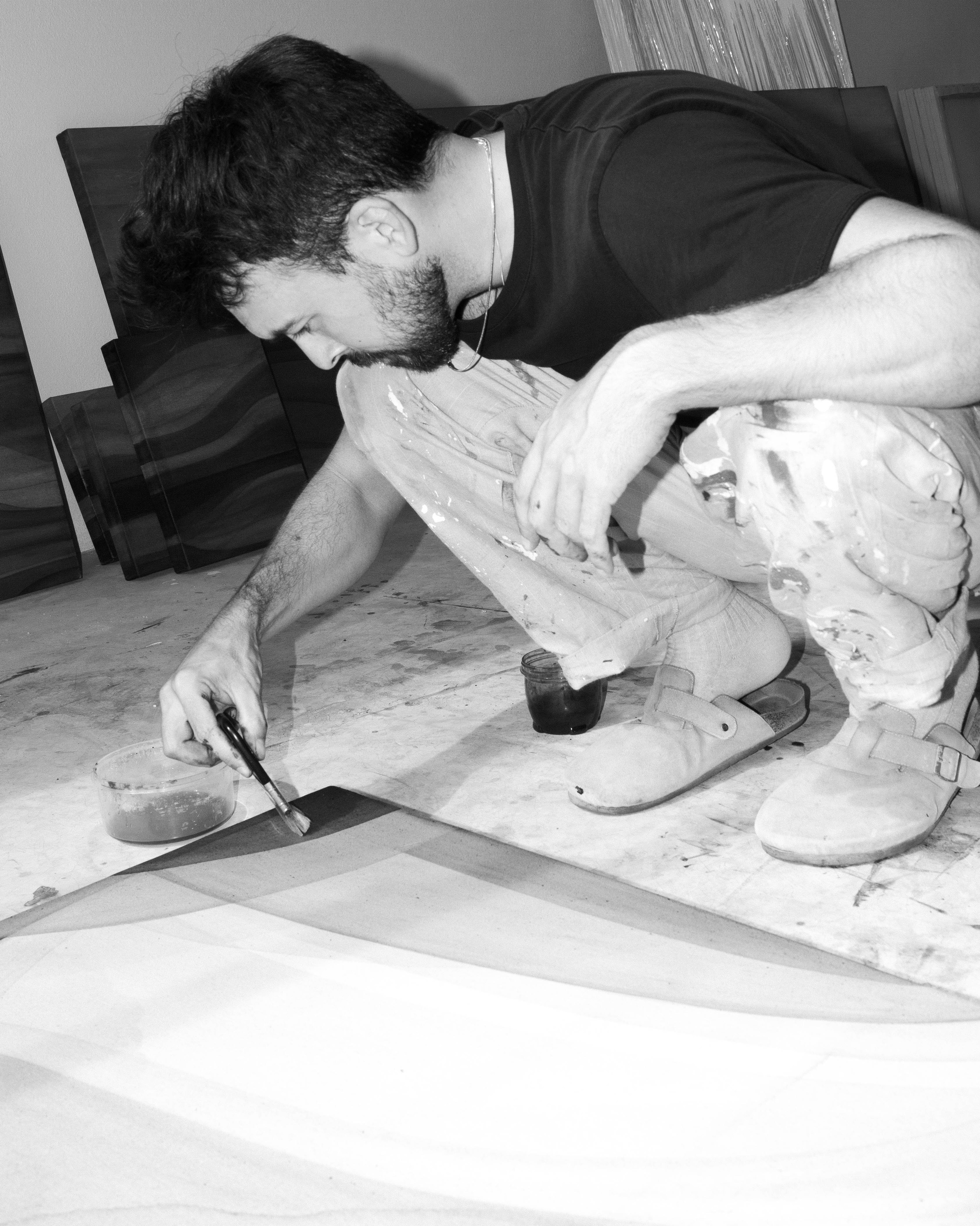 An artist crouching on the studio floor while applying natural pigment to a canvas with a brush.
