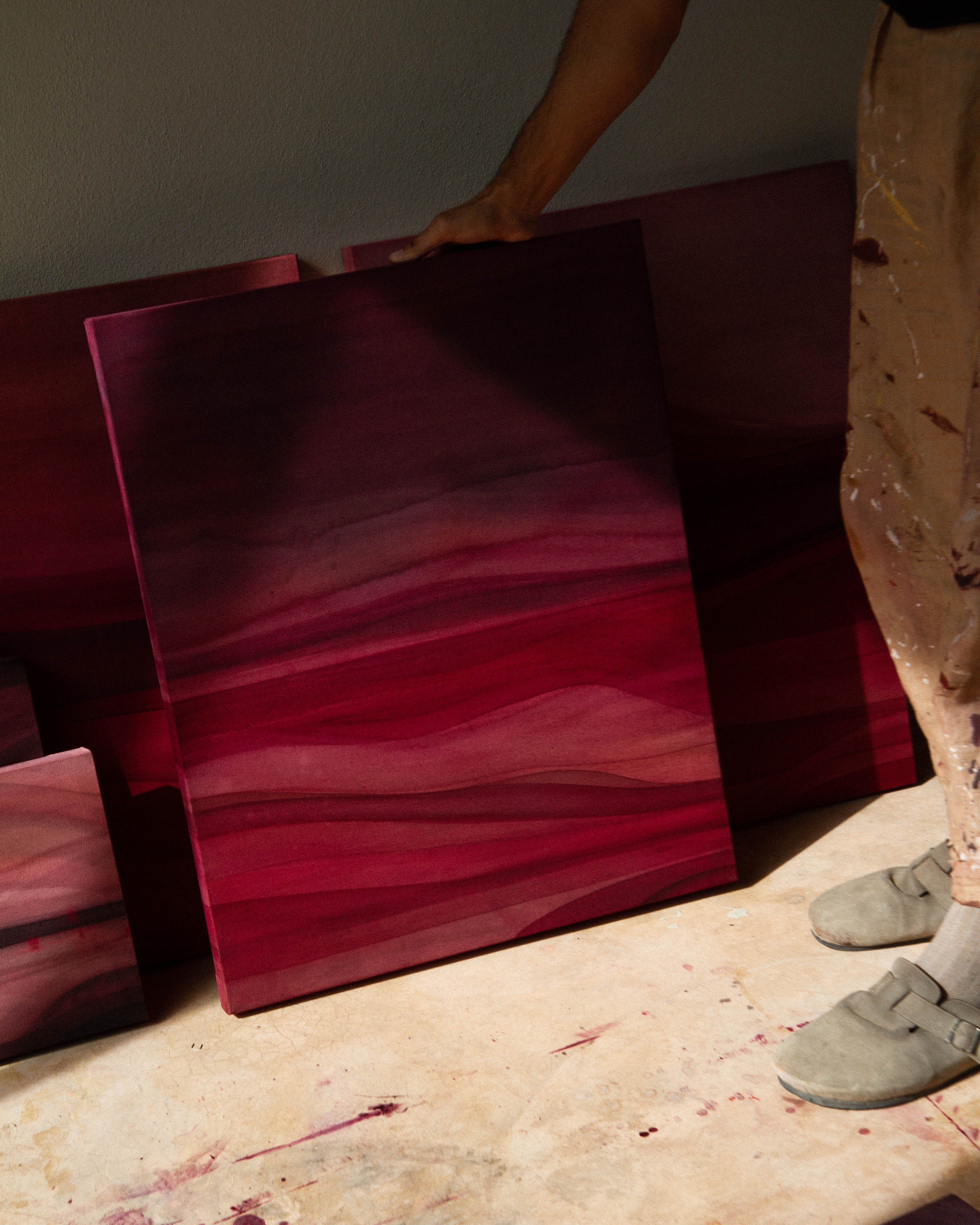 An artist standing in a studio holding an abstract painting beside other works in natural light.