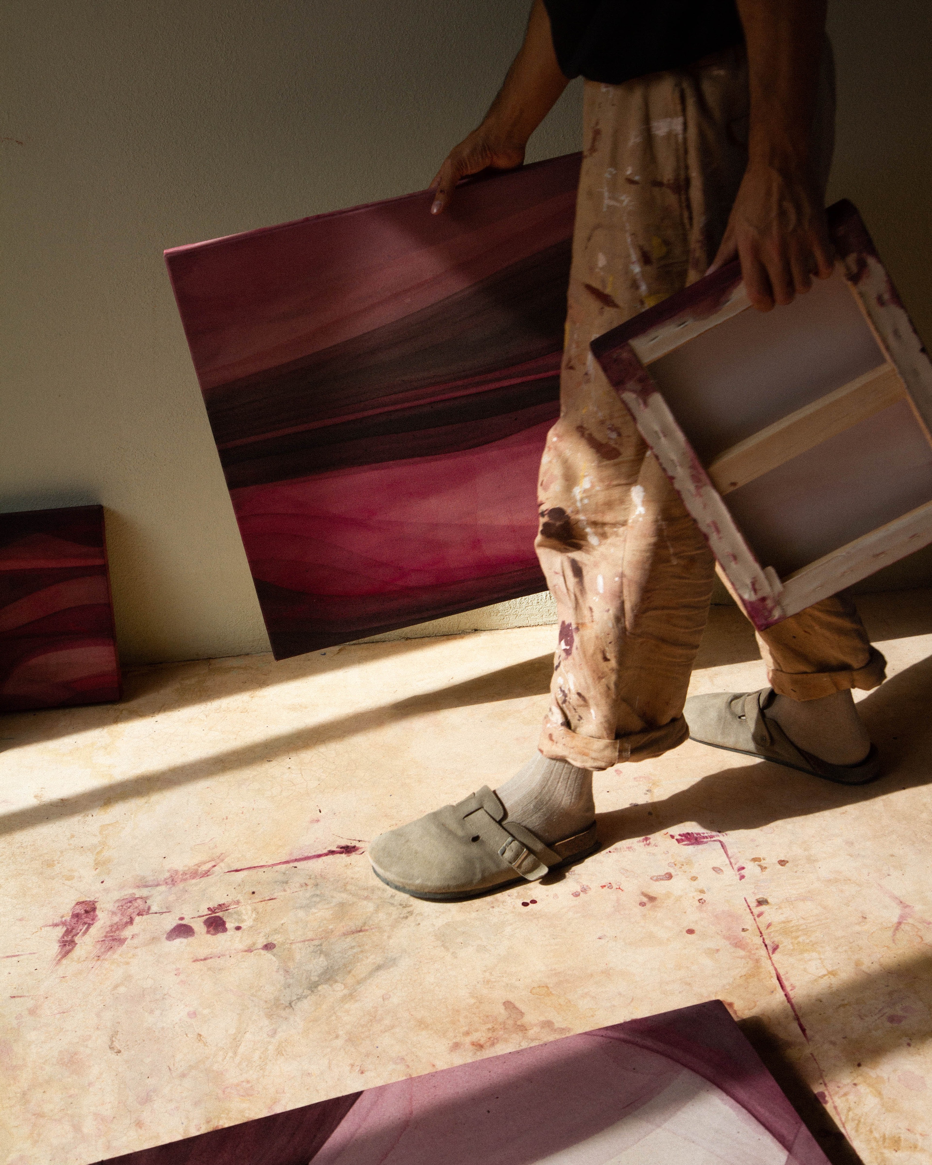 An artist holding an abstract painting while standing on the studio floor beside other works.