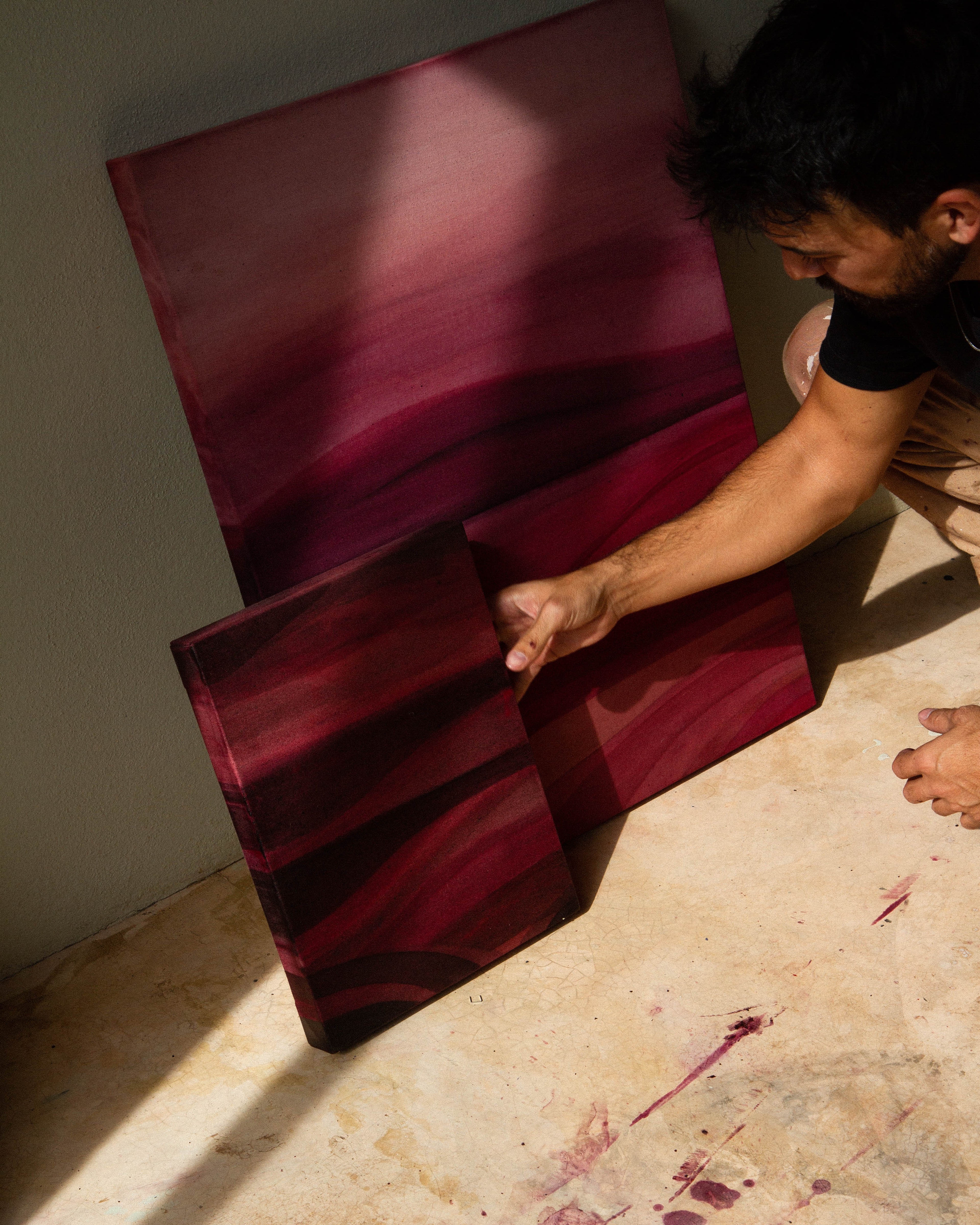 An artist crouching in a studio examining an abstract painting beside other works on the floor.