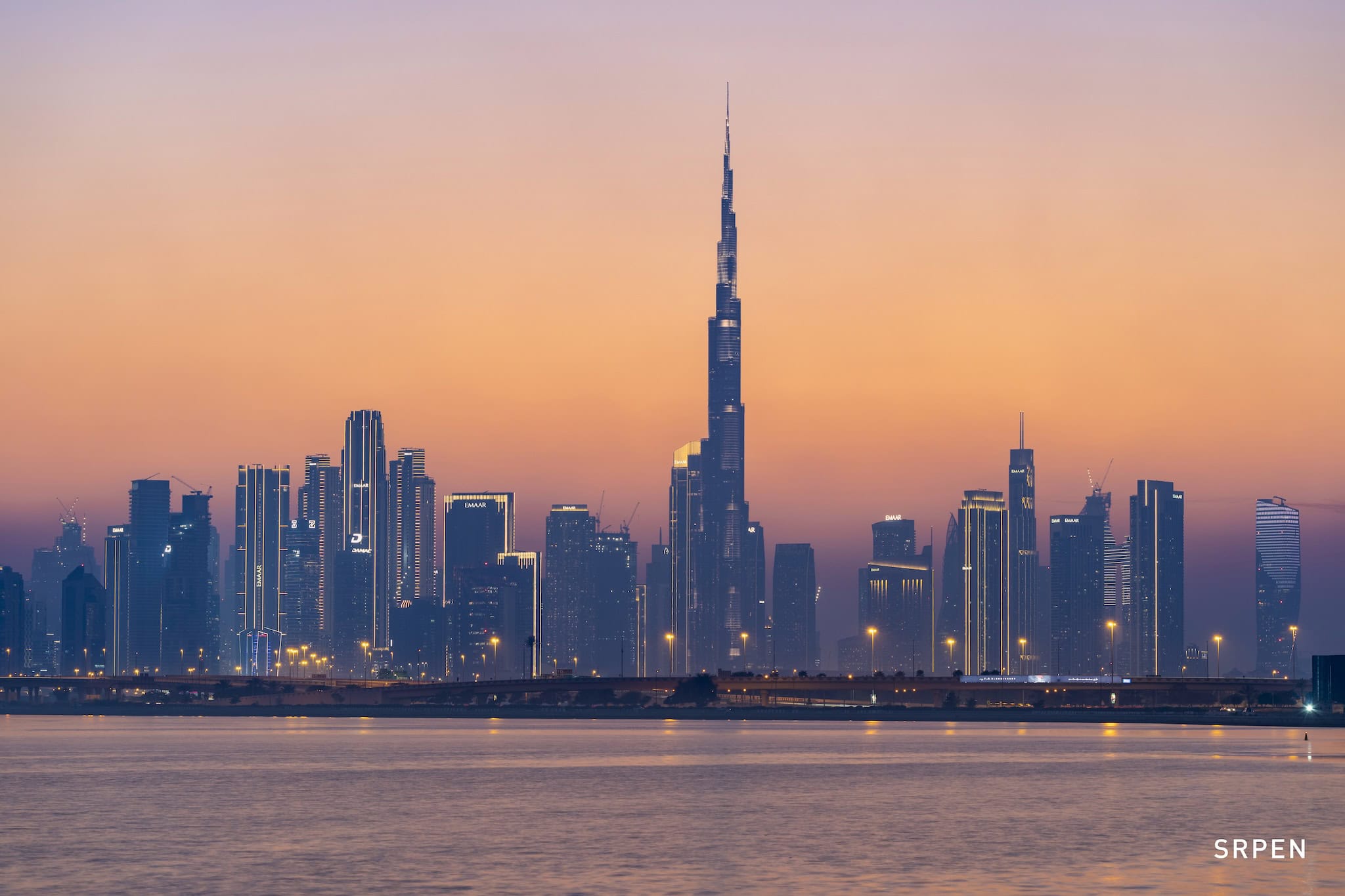 Srpen: panorama města s Burdž Chalífa přes vodní hladinu při západu slunce, Dubaj, Spojené arabské emiráty