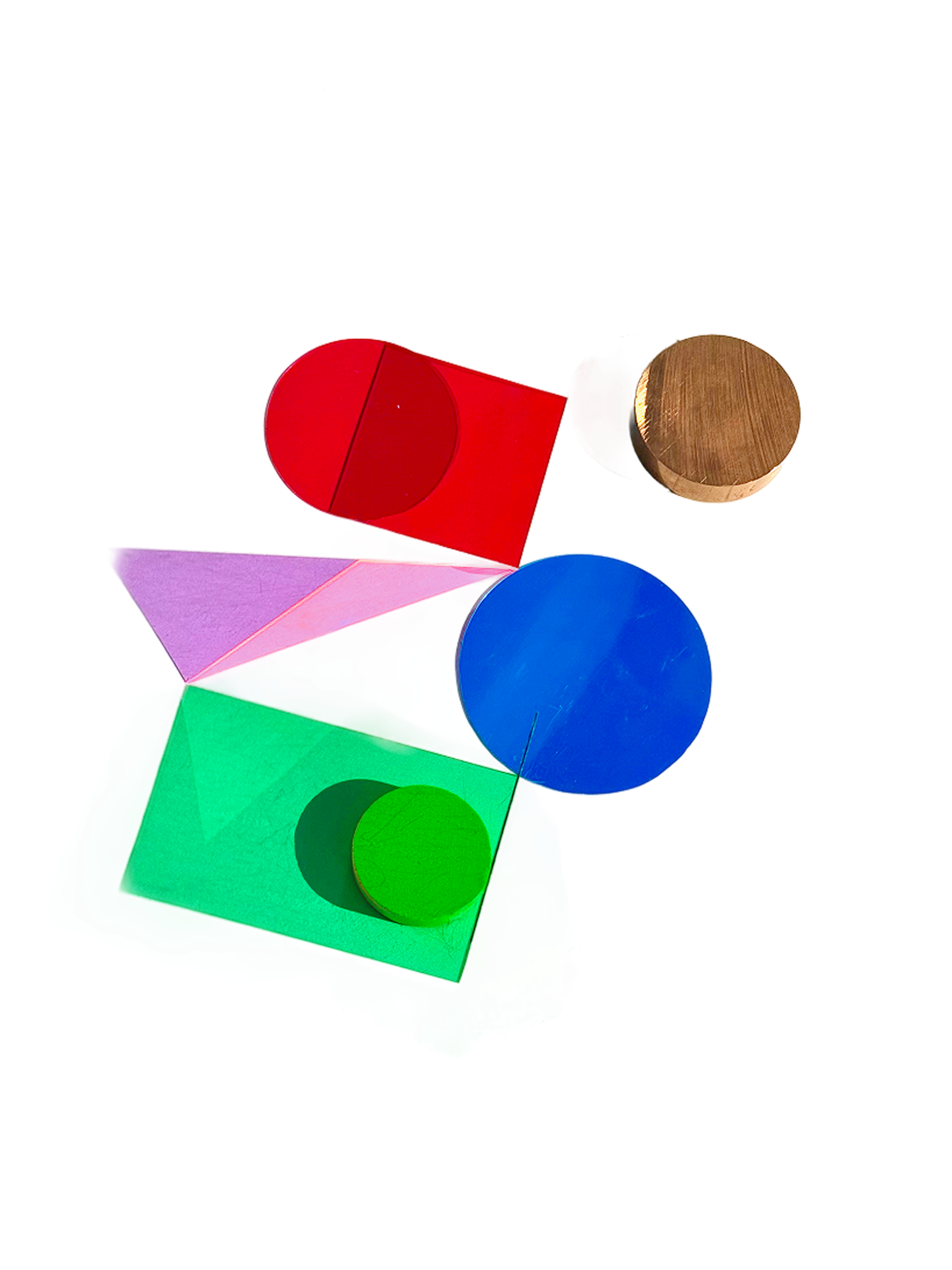 Colorful physical shapes arranged on a white surface, including a red circle, blue disc, green rectangle, purple triangle, wooden cylinder, and green sphere, casting soft shadows