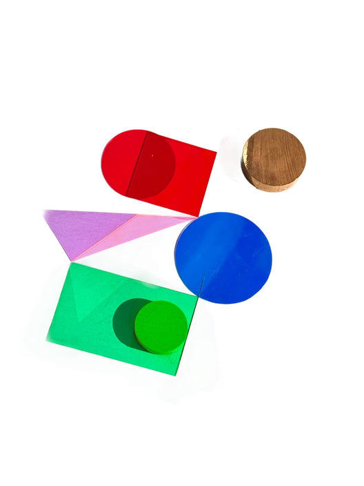 Colorful physical shapes arranged on a white surface, including a red circle, blue disc, green rectangle, purple triangle, wooden cylinder, and green sphere, casting soft shadows