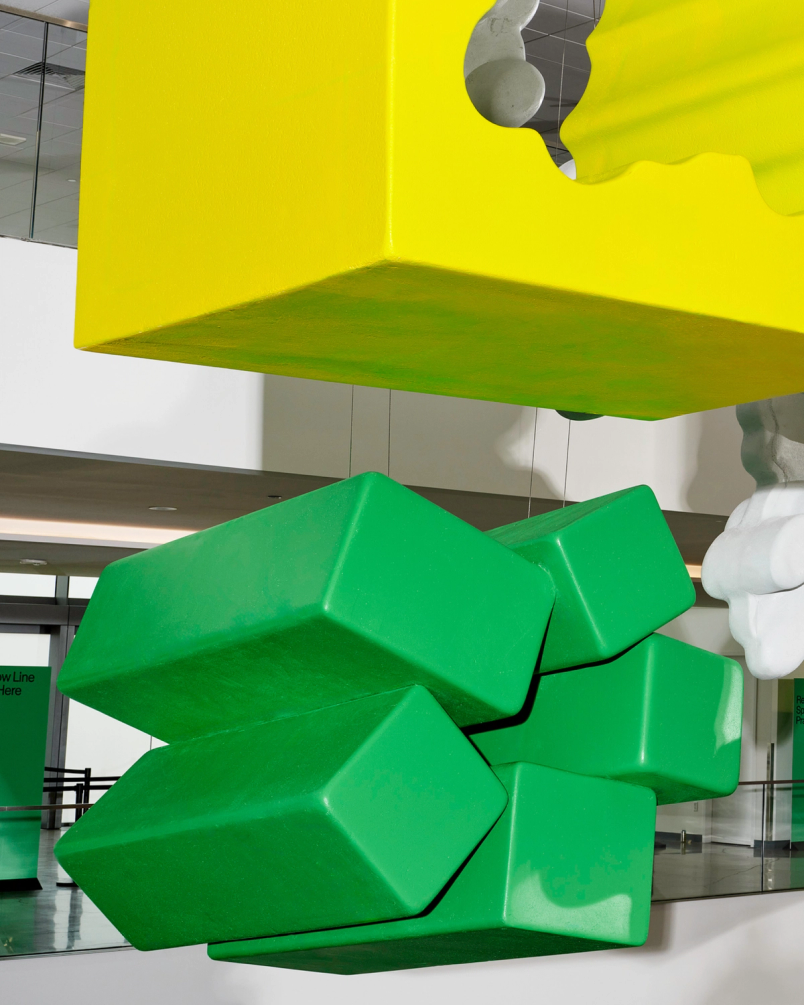 Oversized geometric sculptures in yellow, green, and white hang from the ceiling of a glass-walled atrium.