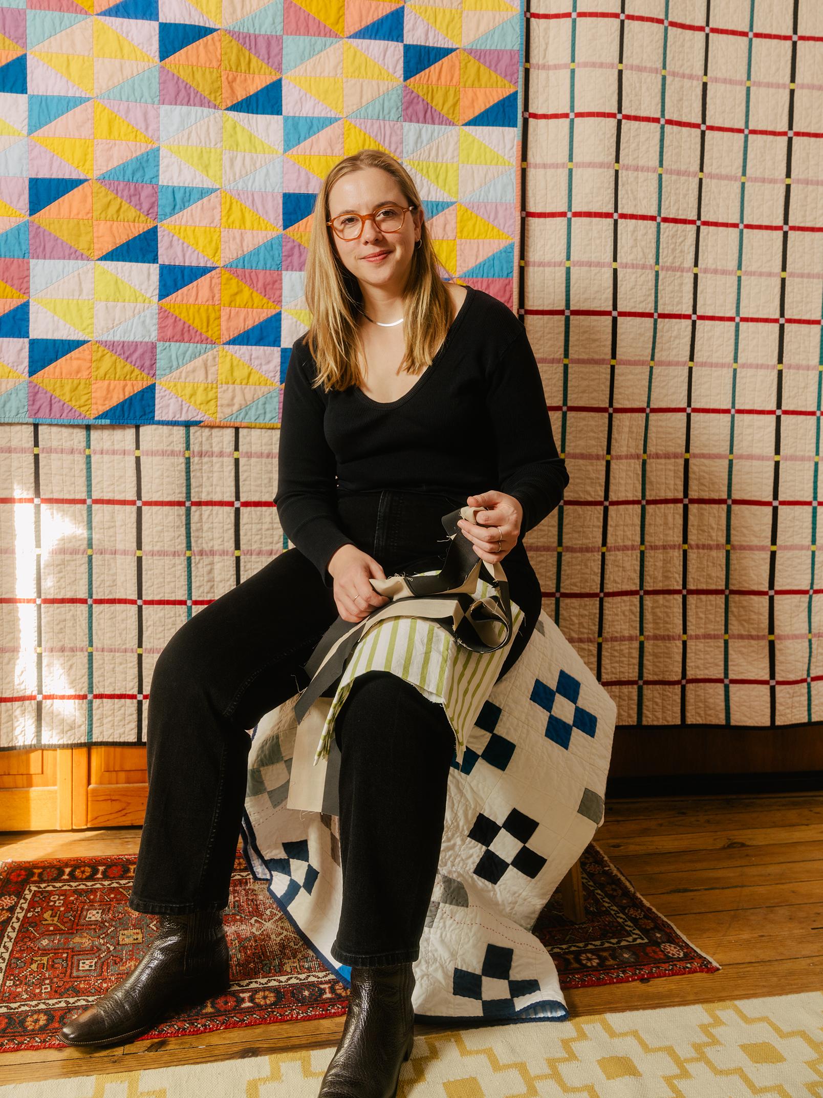 A photograph of Nicole, who has long blonde hair and is wearing red glasses, sitting in front of her quilts